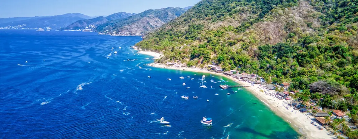 Panoramic aerial view of Las Ánimas Beach in Puerto Vallarta, showcasing the coastline with light sand, turquoise water, lush vegetation on the mountains, and boats anchored in the bay.